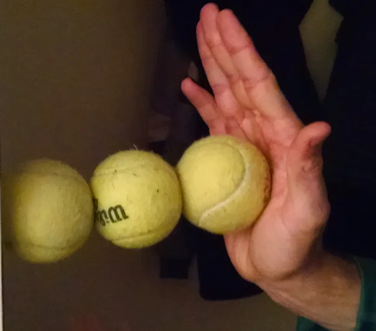Three tennis balls in a line with the one on the left pressed against the wall, and the one on the right pressed against a person's hand.