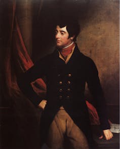 A man with thick dark hair poses in a riding jacket and tan pants, with his fist on one of his hips.