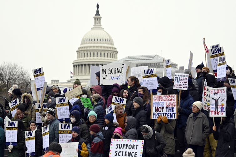 Protectors holding signs reading 'SUPPORT SCIENCE' and 'IN SCIECE WE TRUST,' among others