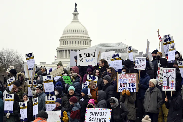 Protectors holding signs reading 'SUPPORT SCIENCE' and 'IN SCIECE WE TRUST,' among others