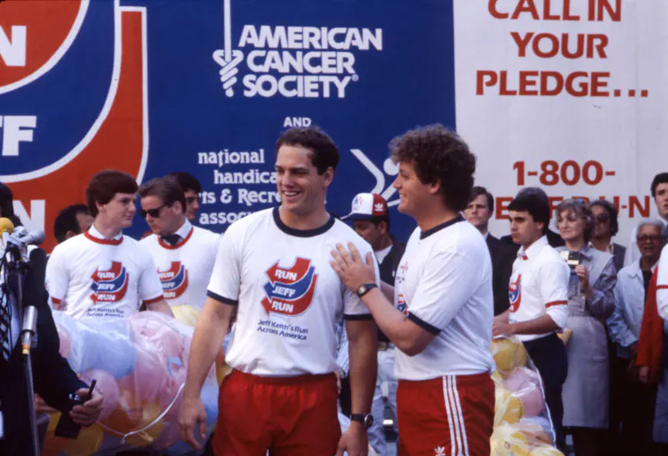 Crowd of people in white T-shirts reading 'RUN JEFF RUN' standing in front of a backdrop of a sign with the American Cancer Society logo and another sign reading 'CALL IN YOUR PLEDGE...'