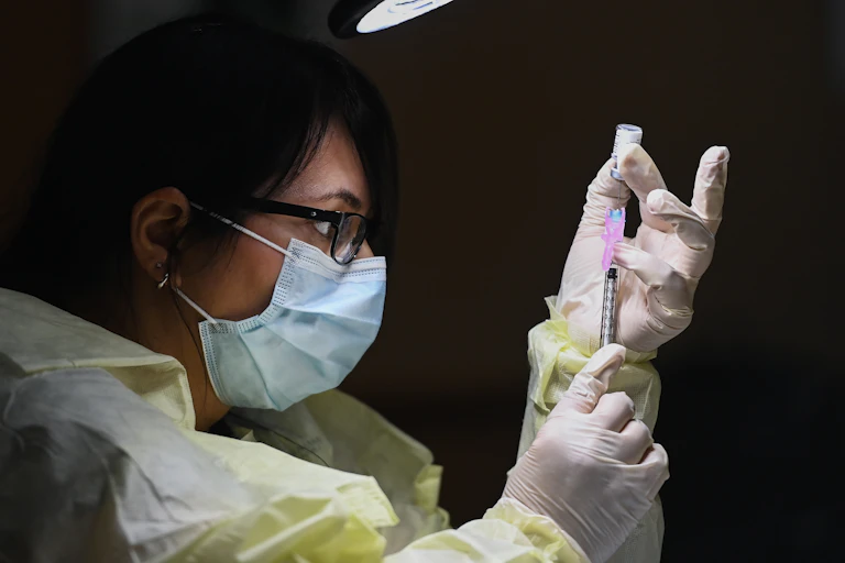 Close-up of a woman in profile in PPE gown, gloves and face mask filling a syringe