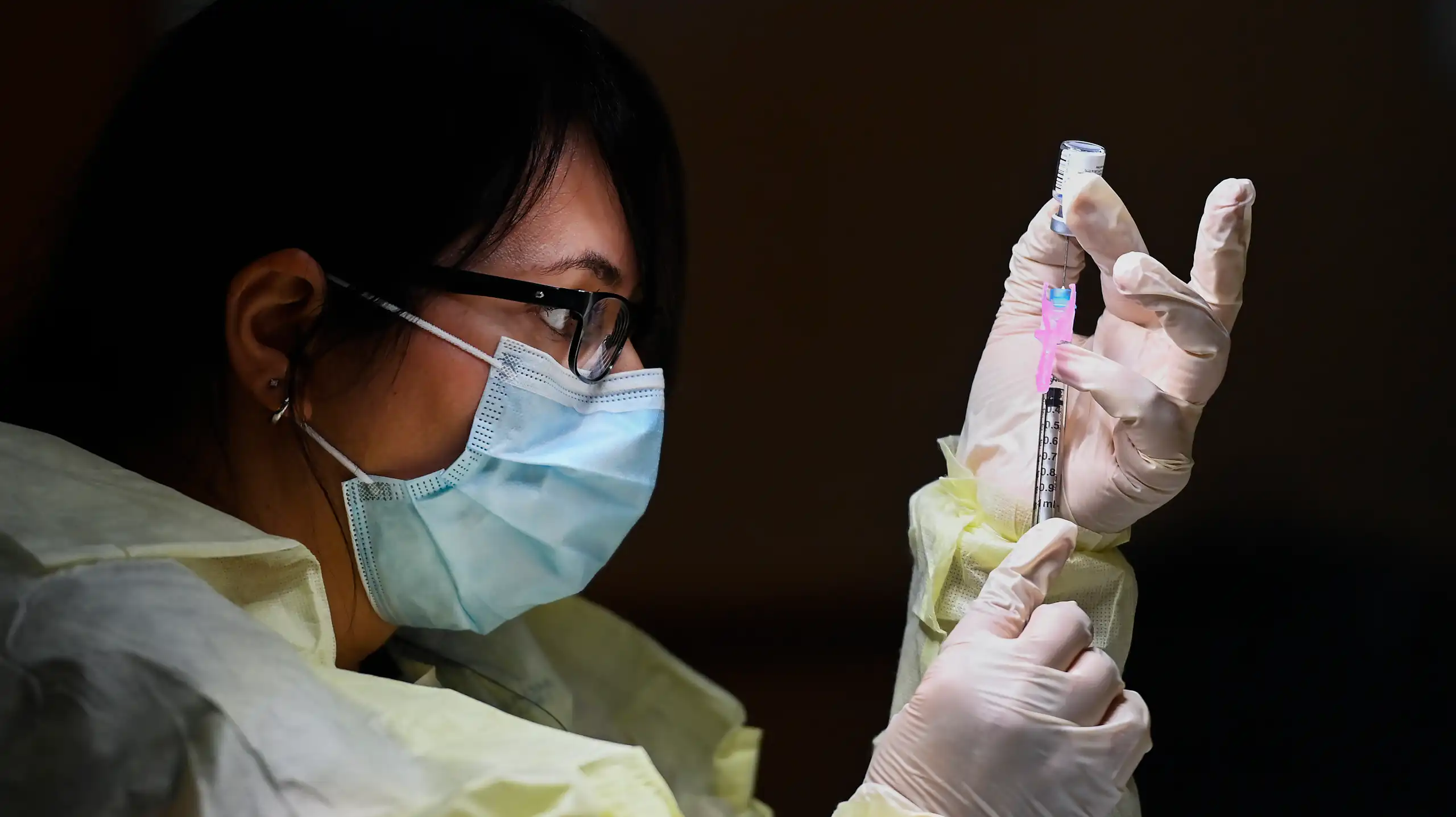 Close-up of a woman in profile in PPE gown, gloves and face mask filling a syringe