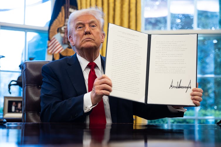 Donald Trump sits at the Resolute Desk and holds up an executive order on auto tariffs, April 2025.