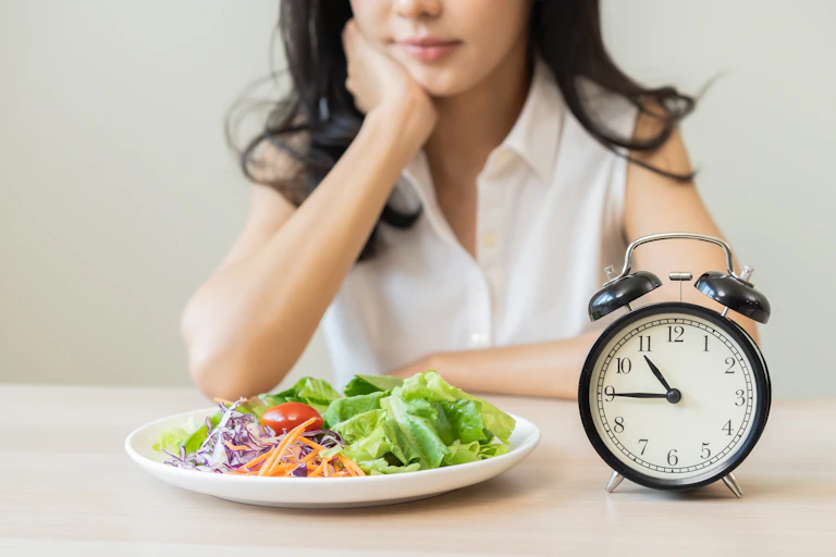A woman sits at a table. In front of her is a plate of a low-carb salad and a black clock.