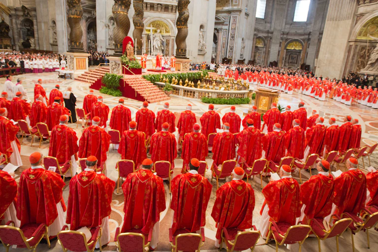 Men dressed in priestly red robes stand in circular rows in an ornate hall around a raised podium.