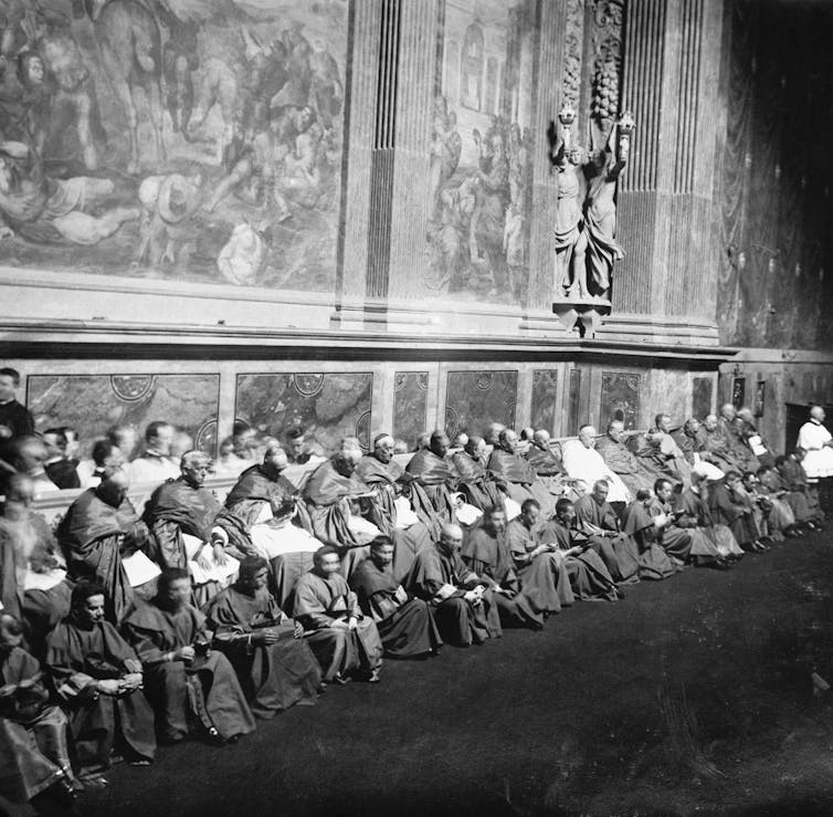 A black-and-white photograph shows men dressed in long robes, seated in rows. On the wall behind them are large frescoes.