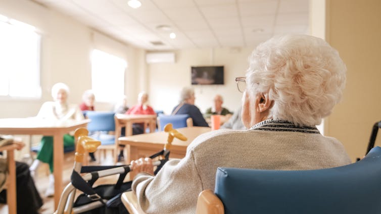 An older woman using a wheelchair, seen from behind entering a room where others are seated at tables