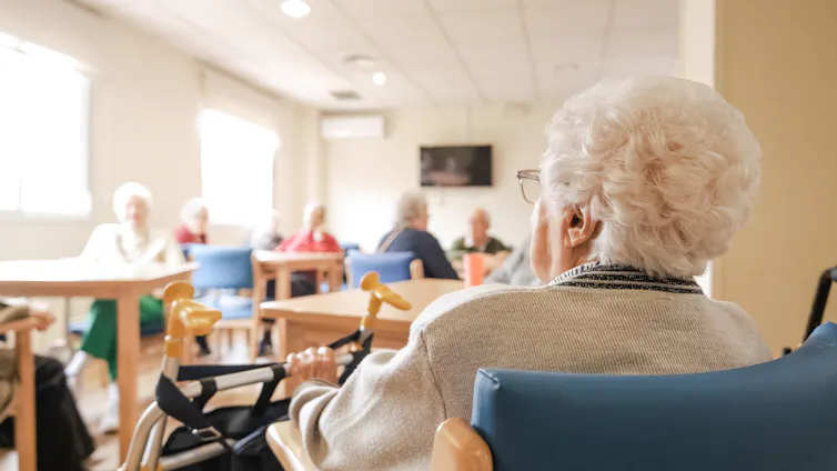 An older woman using a wheelchair, seen from behind entering a room where others are seated at tables