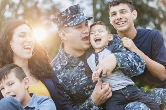 An Hispanic man in the U.S. military, dressed in camouflaged clothes, enjoying time with his wife and children.
