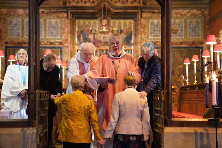 Two women in formal jackets kneel before a clergyman in an orange robe.