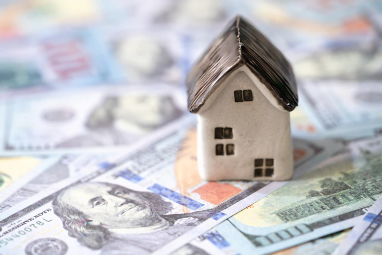 A model home sits on top of several $100 bills spread across a flat surface.
