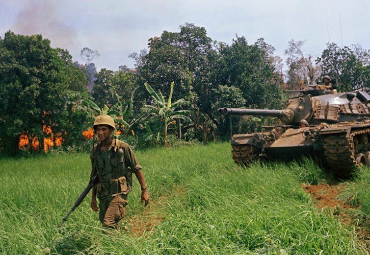 A tank and soldier pass by a burning area of forest.
