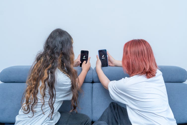 two young women look at their phones while seated on the floor near a blue ottoman