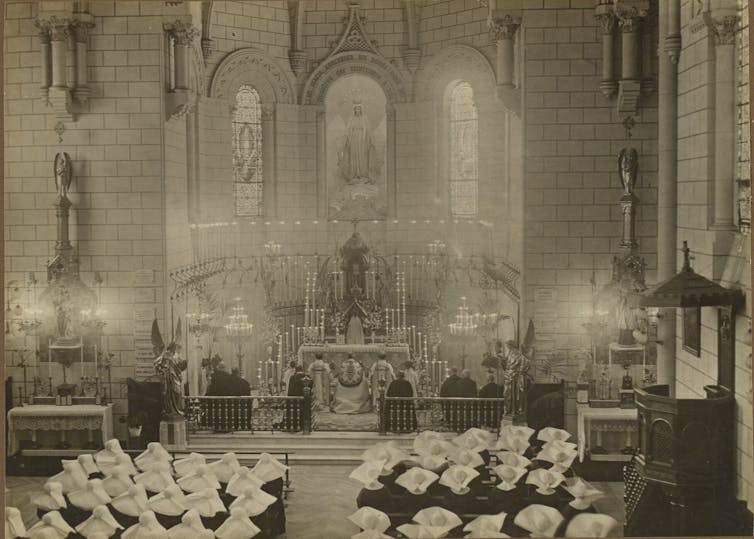 Photograph: The inside of a cathedral during mass. Nuns sit in rows wearing white cornette hats, facing towards the altar where priests and bishops kneel on the ground. Lit candles cover the altar.