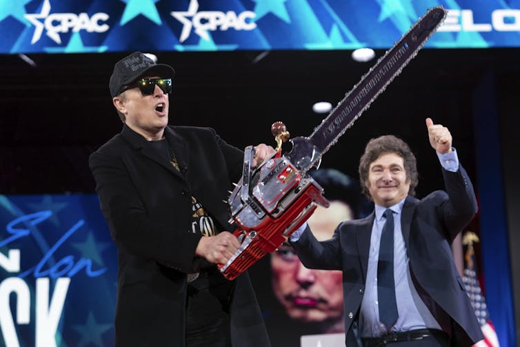 Two men posture on stage at a political rally, Elon Musk wearing sunglasses with a chainsaw in the foreground.