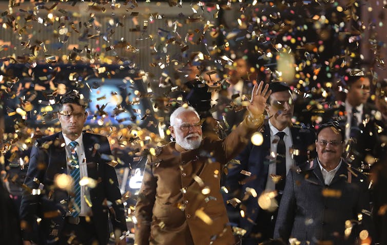 A group of smiling men walk through golden confetti while waving at a crowd.