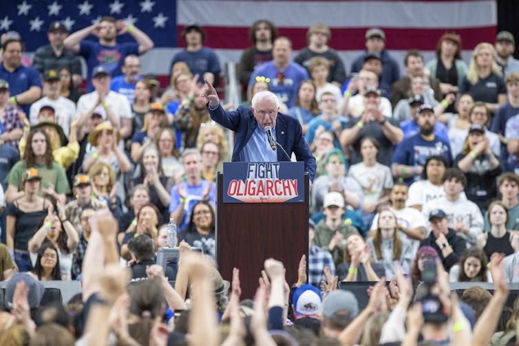 A man at a public rally raises his fist while speaking at a lectern.