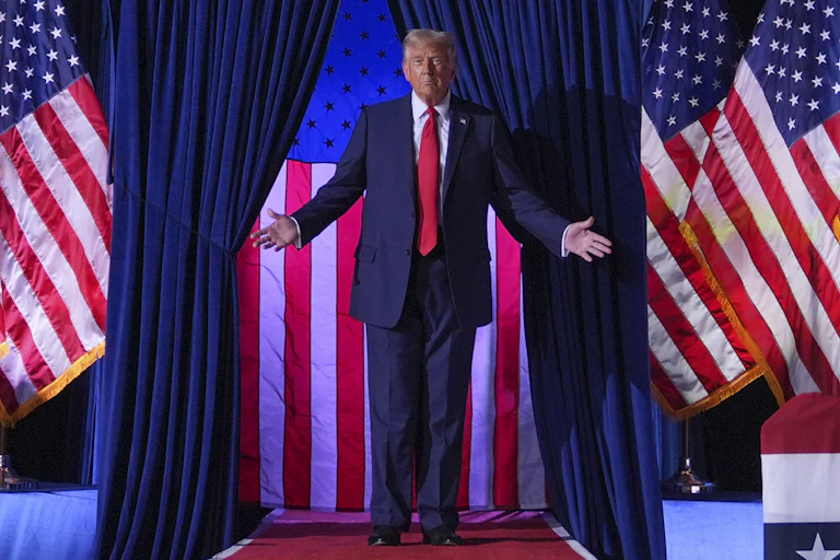 A man in a suit spreads his arms wide while standing in front of US flags.