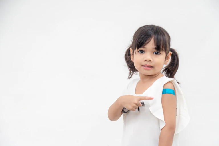 A smiling young girl points to a small blue plaster on her upper arm, indicating that she has just had a vaccine.