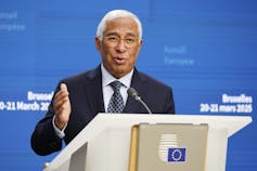 A tanned man with white hair speaks at a podium.