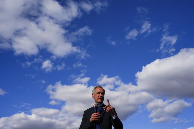 A man with short grey hair speaks into a microphone with a large blue sky as a backdrop.