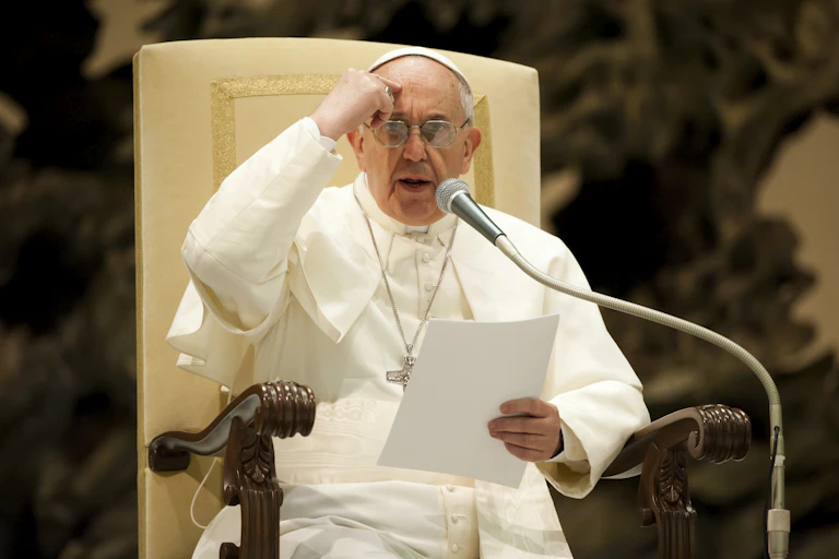 A man in a white robe speaks into a microphone while seated