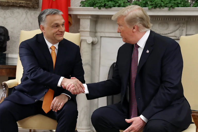 Two men in dark suits shake hands while sitting in chairs.
