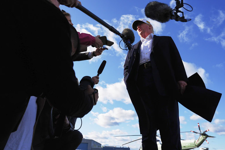 A rotund man in a white ball cap is surrounded by microphones and reporters on an airport tarmac.