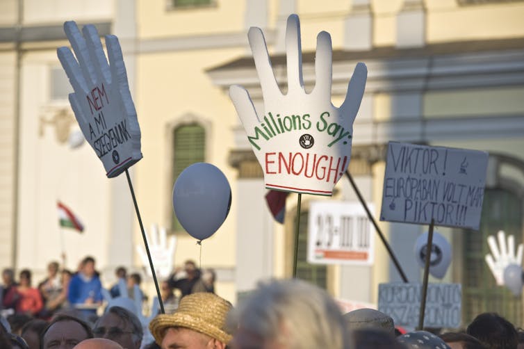 A crowd of protesters in front of a big white building, holding large signs in the shape of hands , with one saying 'Millions say ENOUGH.'