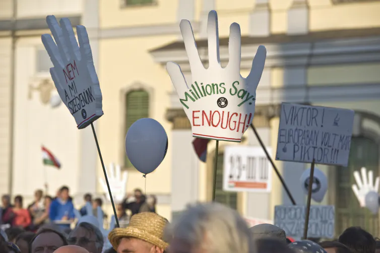 A crowd of protesters in front of a big white building, holding large signs in the shape of hands , with one saying 'Millions say ENOUGH.'