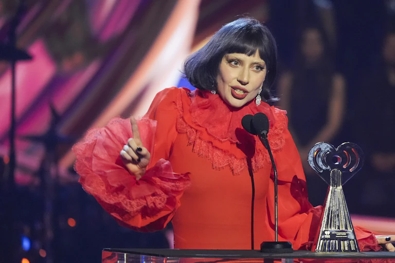 A woman in a red blouse at a podium speaking into a microphone holding up her finger.