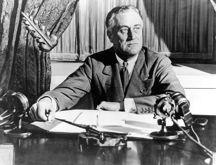 A black and white photo shows a grey-haired man wearing glasses at a desk with a microphone in front of him.
