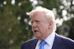 An elderly man with fluffy blond hair looks tired as he speaks to reporters outside.