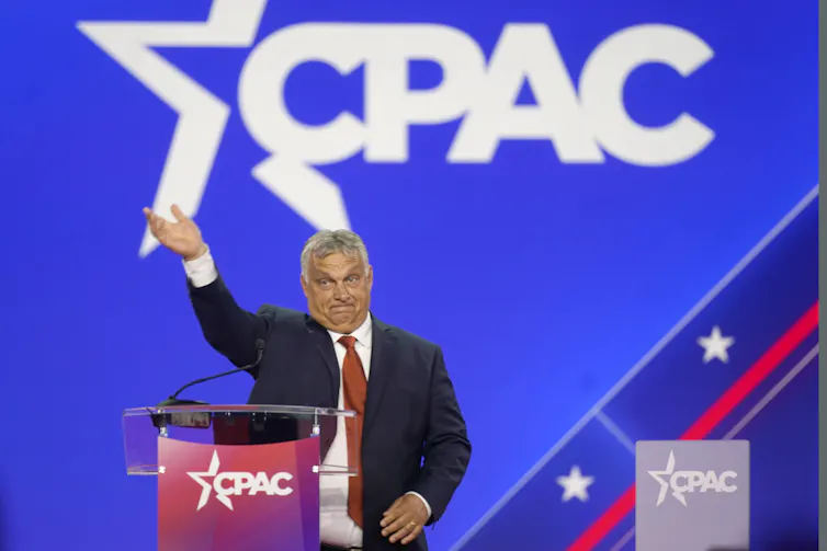 A man in a suit waving from a stage with the logo of the organization known as CPAC behind him.