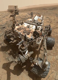 The Curiosity rover on the dusty surface of Mars. The rover has six thick wheels and multiple scientific instruments and cameras.