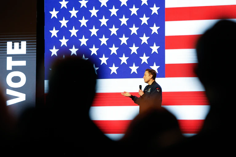 A white man speaks on a stage to a crowd of people in front of a giant projection of an American flag