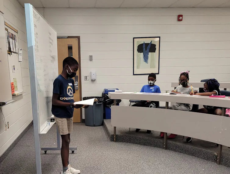 A Black student presenting in a classroom at the front as other students facing them listen.