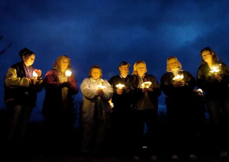 Seven people standing in a row hold candlelights at dusk.