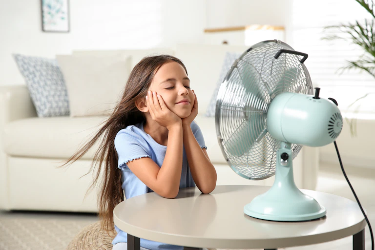Little girl enjoying the air flow from an electric fan at home during summer.