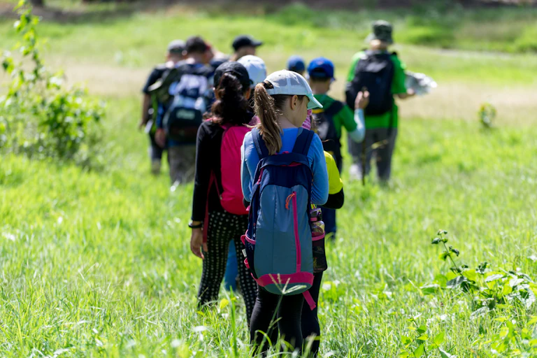 A group of young children in backpacks walk in a green field.