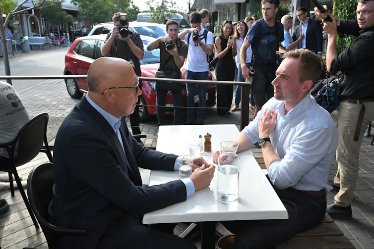 Two men have a coffee and a table in front of a large press pack