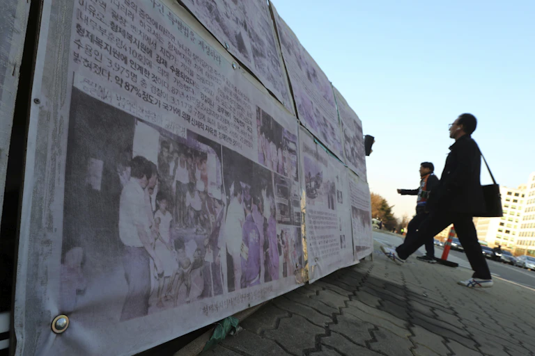 In this April 2, 2019, photo, people walk past a photo of guards unloading children from a truck at the Brothers Home in Busan, as it is displayed in front of National Assembly in Seoul, South Korea.