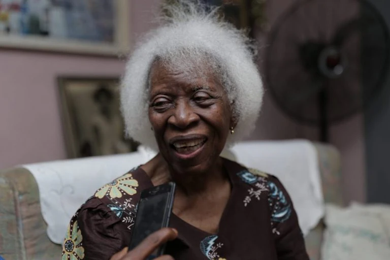 A white-haired woman speaking to someone who is holding a phone.