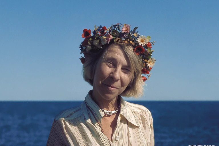An older woman with a flower garland on her head smiling, the sea in the background.