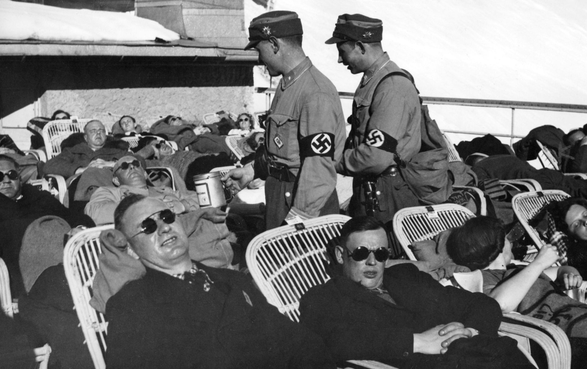 A black and white photo of tourists lying on chairs in the sun while two Nazis pass through the crowd