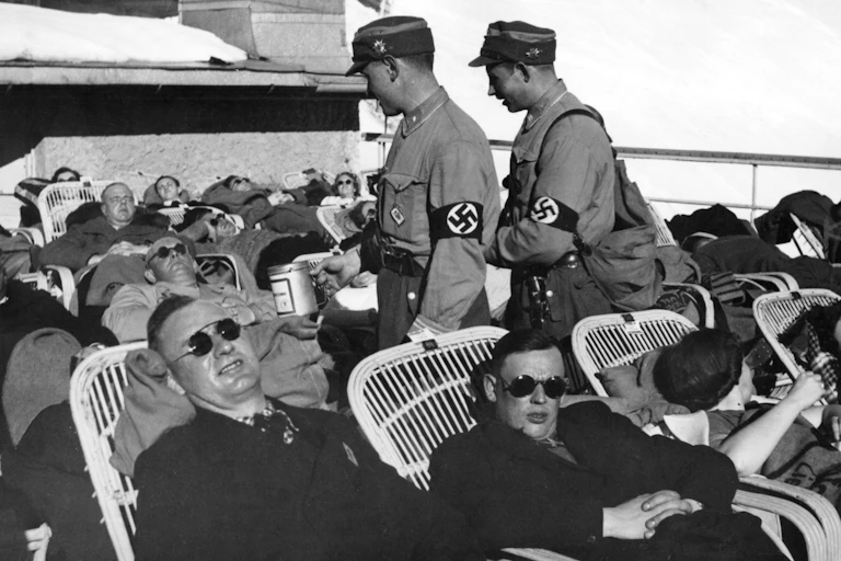 A black and white photo of tourists lying on chairs in the sun while two Nazis pass through the crowd