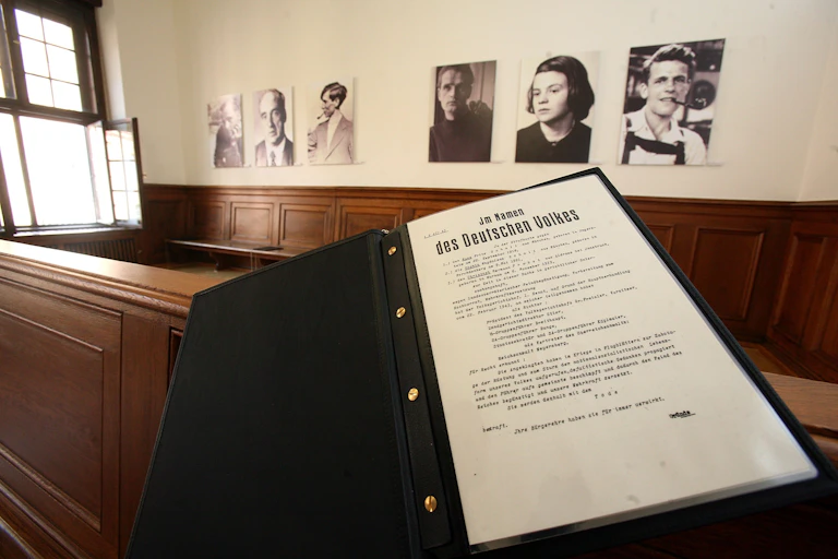 An austere-looking room has six photographs of men and women displayed on the wall, while in the foreground lies a large open folder with a document.