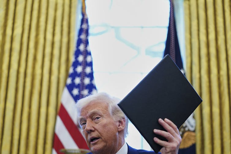 El hombre mayor con el cabello de aleteo blanco mantiene el documento. Las cortinas de oro y las banderas americanas están detrás de él.