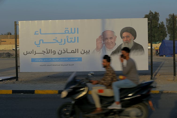 A poster depicts a man in a white priestly robe standing beside another man wearing a black turban, while two men on a motorbike pass by.